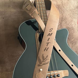 May include: A light brown leather guitar strap with the name "SMITH" vertically engraved. The strap features a detailed illustration of two flying ducks and is draped over a black acoustic guitar.