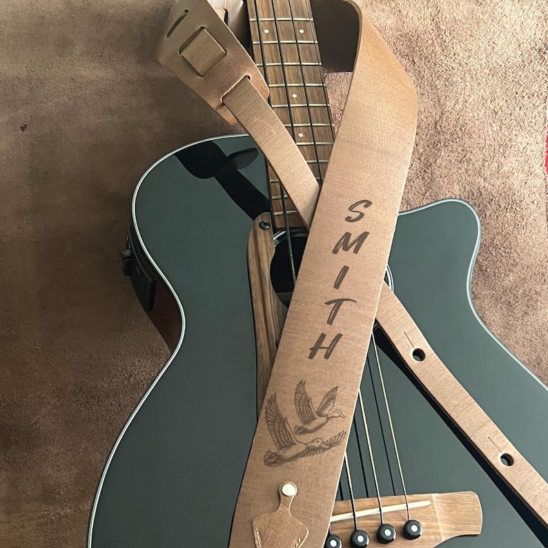 May include: A light brown leather guitar strap with the name "SMITH" vertically engraved. The strap features a detailed illustration of two flying ducks and is draped over a black acoustic guitar.