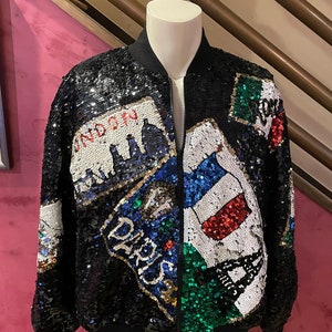 Könnte beinhalten: Eine schwarze Bomberjacke mit einem Pailletten-Design, das verschiedene Städtenamen und Flaggen zeigt, darunter London, Paris und Italien.