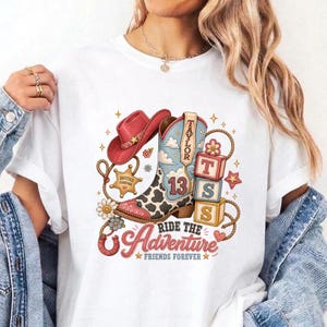 K&ouml;nnte beinhalten: Ein wei&szlig;es T-Shirt mit einem Grafikdruck eines rosa Cowboyhuts, einem Cowboystiefel mit Kuhfleckenmuster und der Zahl 13 sowie drei gestapelten Bl&ouml;cken mit den Buchstaben T, S, S. Der Text lautet Taylor, Ride the Adventure und Besties Forever.