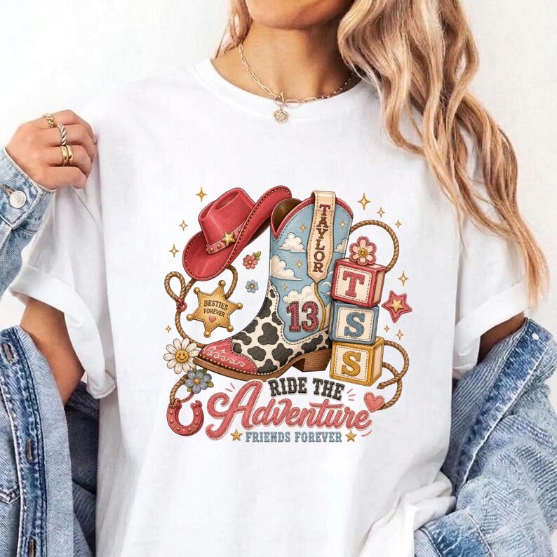 K&ouml;nnte beinhalten: Ein wei&szlig;es T-Shirt mit einem Grafikdruck eines rosa Cowboyhuts, einem Cowboystiefel mit Kuhfleckenmuster und der Zahl 13 sowie drei gestapelten Bl&ouml;cken mit den Buchstaben T, S, S. Der Text lautet Taylor, Ride the Adventure und Besties Forever.