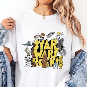 Puede incluir: Una camiseta blanca con un estampado gráfico que incluye el texto amarillo STAR WARS ROCKS! rodeado de ilustraciones de personajes y vehículos de la franquicia Star Wars, incluyendo a Darth Vader, Chewbacca y R2-D2.