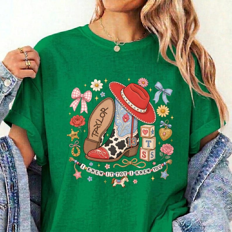 May include: A green t-shirt featuring a graphic print of a red cowboy hat, a cow-print boot with the name Taylor, and alphabet blocks. The design includes flowers, bows, and the text I KNEW IT TOY I KNEW YOU arranged in a circular border.