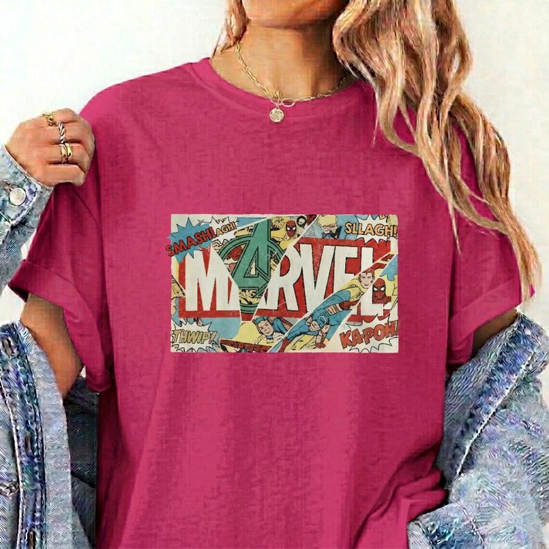 Puede incluir: Una camiseta de manga corta color magenta con un estampado gr&aacute;fico rectangular en el pecho. El gr&aacute;fico muestra la palabra MARVEL en letras blancas grandes, superpuesta sobre un collage de paneles de c&oacute;mics antiguos con palabras de acci&oacute;n.