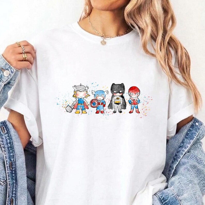 May include: A white short-sleeved t-shirt featuring a colourful watercolour-style graphic of four chibi-proportioned superheroes standing in a row, including characters resembling Thor, Captain America, Batman, and Spider-Man with paint splatters.