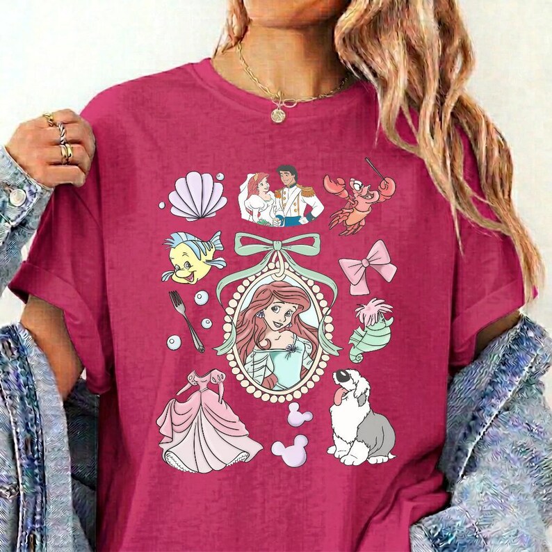 May include: A magenta short-sleeved t-shirt featuring a central oval frame containing a red-haired character, surrounded by various illustrations including a seashell, a fork, a pink bow, a green sea creature, a dog, and a pink dress.
