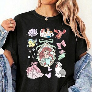May include: A black short-sleeved t-shirt featuring a central oval frame containing a red-haired character, surrounded by various illustrations including a seashell, a crab, a fork, a pink dress, a dog, and a couple in a wedding outfit.