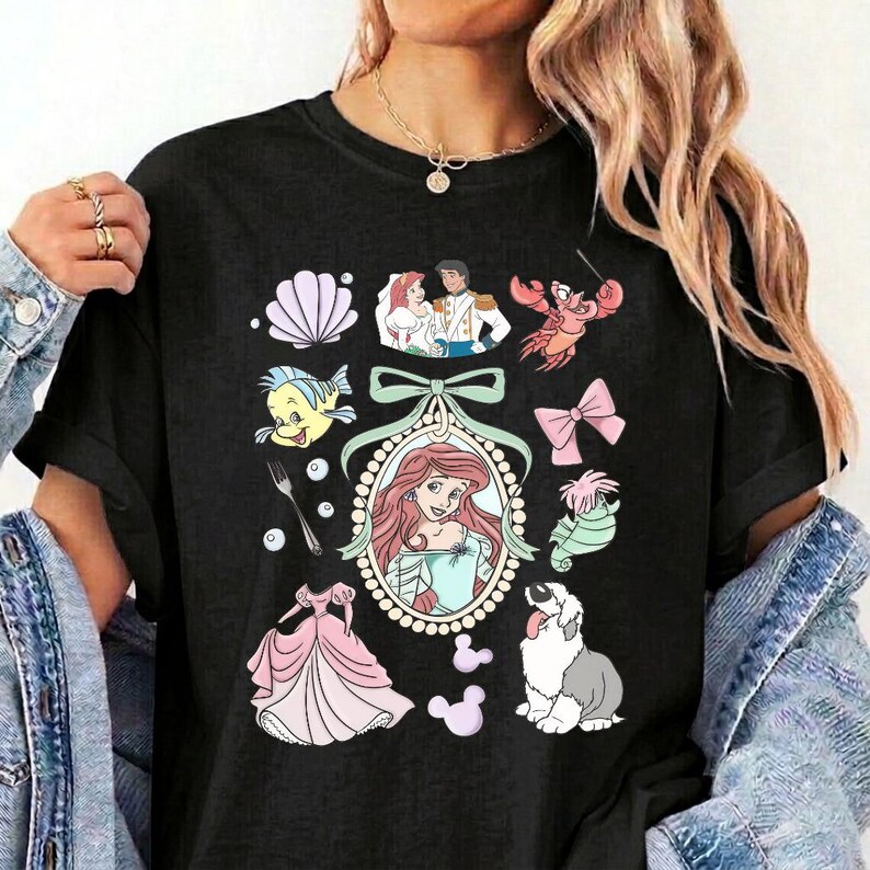 May include: A black short-sleeved t-shirt featuring a central oval frame containing a red-haired character, surrounded by various illustrations including a seashell, a crab, a fork, a pink dress, a dog, and a couple in a wedding outfit.