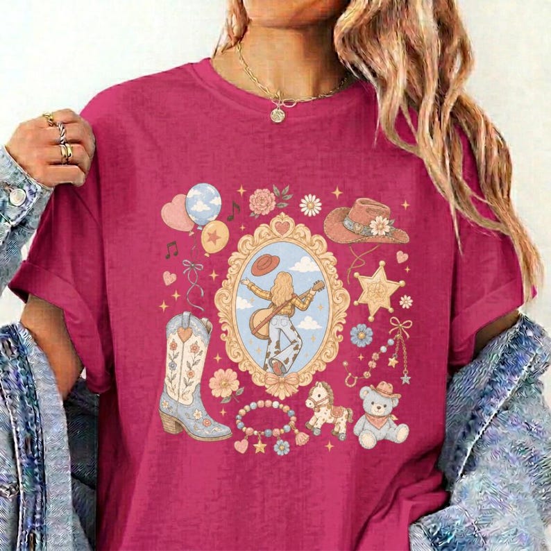 May include: A magenta t-shirt featuring a central ornate gold frame containing an illustration of a person playing a guitar. Surrounding the frame are graphics of a cowboy boot, a hat, a teddy bear, a rocking horse, balloons, and flowers.
