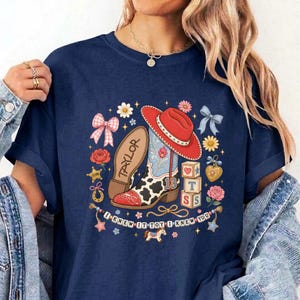 May include: A navy blue t-shirt featuring a graphic print of a red cowboy hat, a cow-print boot with the name Taylor, and alphabet blocks. The design includes flowers, bows, stars, and a banner with the text I KNEW IT TOY I KNEW YOU.