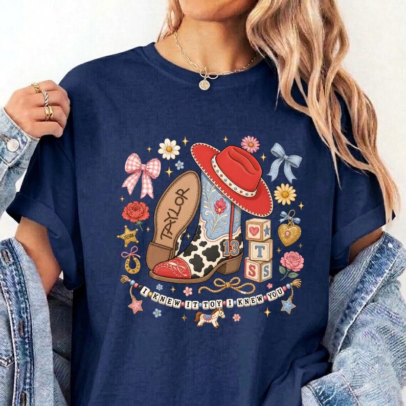 May include: A navy blue t-shirt featuring a graphic print of a red cowboy hat, a cow-print boot with the name Taylor, and alphabet blocks. The design includes flowers, bows, stars, and a banner with the text I KNEW IT TOY I KNEW YOU.