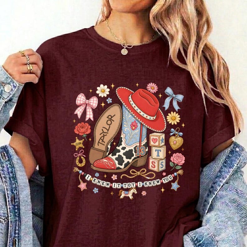 May include: A maroon t-shirt featuring a graphic print of a red cowboy hat, a cow-print boot with the name Taylor, and building blocks with the number 13. The design includes flowers, bows, and the text I KNEW IT TOY I KNEW YOU.