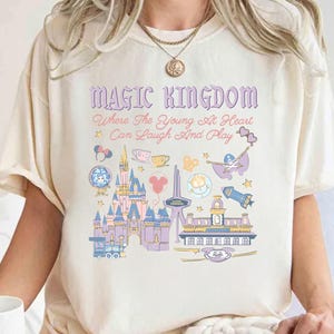 May include: Cream-colored t-shirt with a colorful graphic of a castle, train, and other theme park icons. The text "MAGIC KINGDOM" and a quote are printed above the graphic. The design is in shades of purple, blue, pink, and yellow.