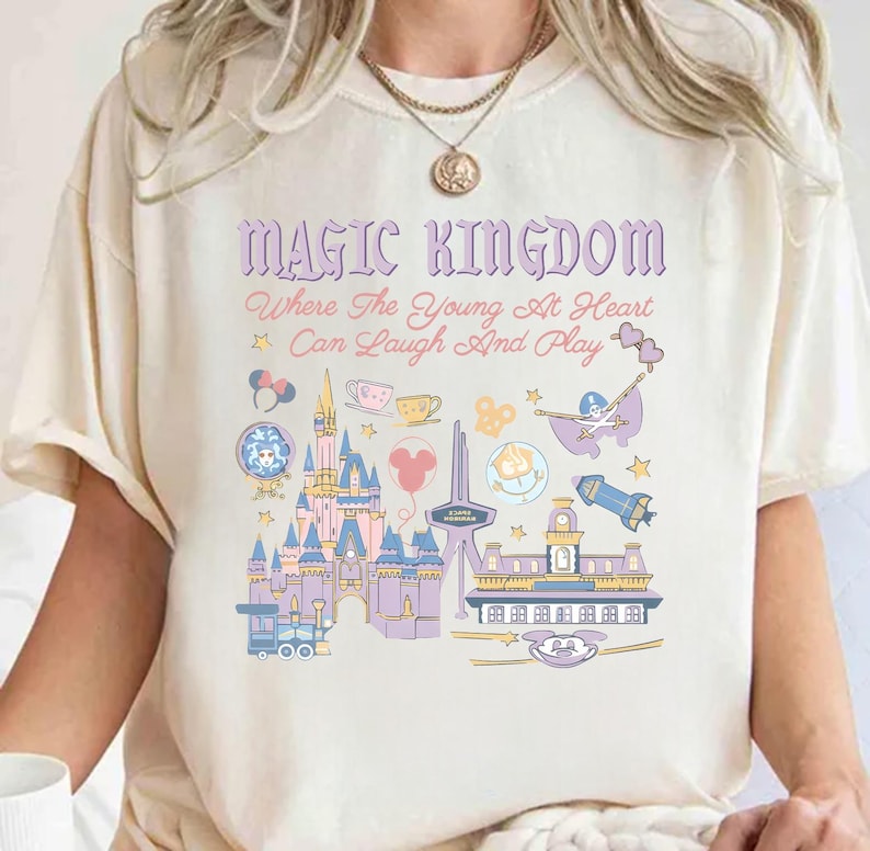 May include: Cream-colored t-shirt with a colorful graphic of a castle, train, and other theme park icons. The text "MAGIC KINGDOM" and a quote are printed above the graphic. The design is in shades of purple, blue, pink, and yellow.