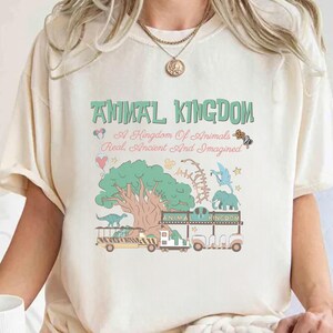 May include: Cream-colored t-shirt with a whimsical "Animal Kingdom" design in green, pink, and blue. The graphic includes a tree, dinosaurs, an elephant, and the text "A Kingdom Of Animals Real, Ancient And Imagined."