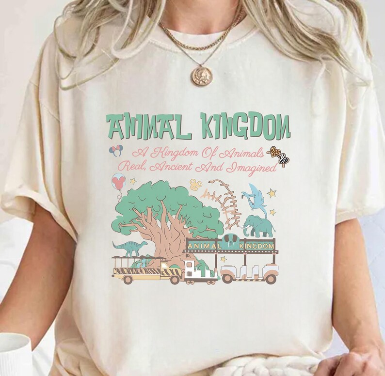 May include: Cream-colored t-shirt with a whimsical "Animal Kingdom" design in green, pink, and blue. The graphic includes a tree, dinosaurs, an elephant, and the text "A Kingdom Of Animals Real, Ancient And Imagined."