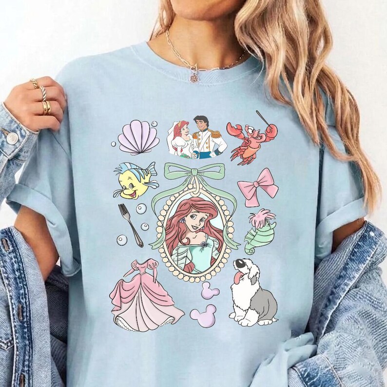 May include: A light blue t-shirt featuring a graphic print of characters and icons from The Little Mermaid. The design includes Ariel in a central oval frame, a fork, a seashell, a crab, a seahorse, a dog, a pink dress, and a bow.