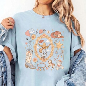 May include: A light blue t-shirt featuring a central graphic print of a person playing a guitar inside an ornate gold frame, surrounded by illustrations of a cowboy boot, a teddy bear, a rocking horse, balloons, a star badge, and flowers.