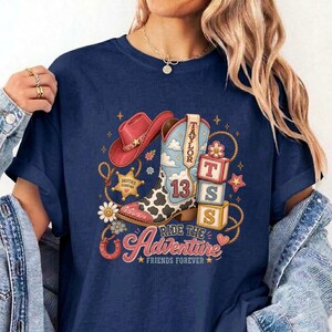K&ouml;nnte beinhalten: Ein marineblaues T-Shirt mit einem Grafikdruck, der einen rosa Cowboyhut, einen Cowboystiefel mit Kuhfleckenmuster und der Zahl 13 sowie drei gestapelte Bl&ouml;cke mit den Buchstaben T, S und S zeigt. Das Design enth&auml;lt den Text Taylor, Ride the Adventure und Friends Forever.