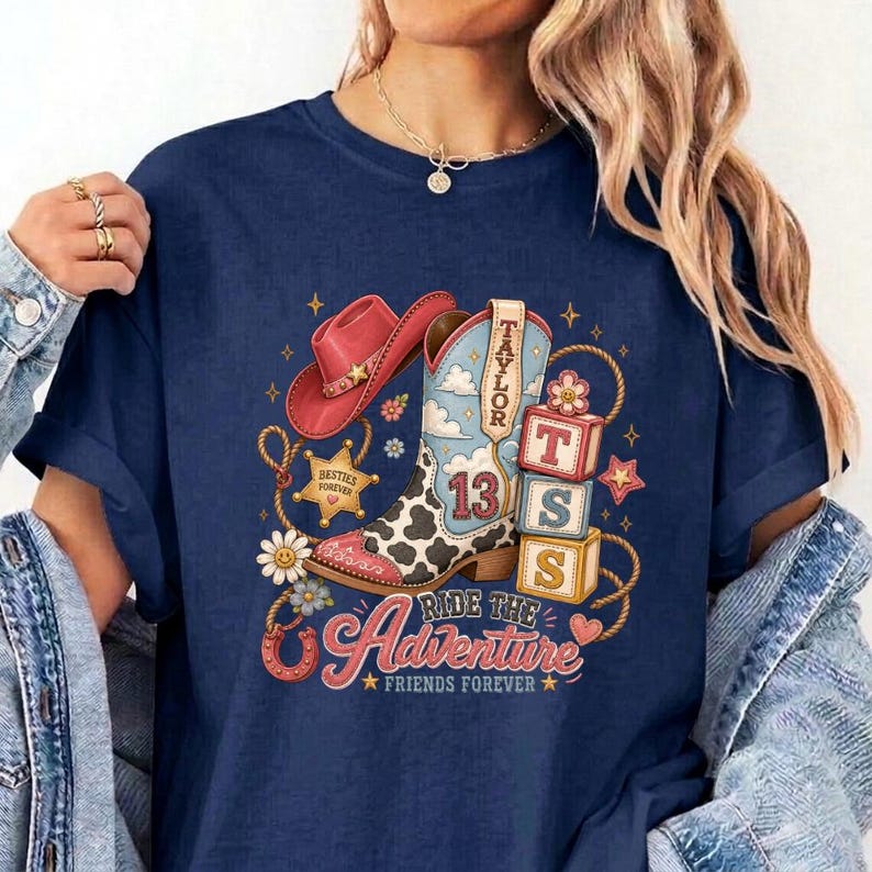 K&ouml;nnte beinhalten: Ein marineblaues T-Shirt mit einem Grafikdruck, der einen rosa Cowboyhut, einen Cowboystiefel mit Kuhfleckenmuster und der Zahl 13 sowie drei gestapelte Bl&ouml;cke mit den Buchstaben T, S und S zeigt. Das Design enth&auml;lt den Text Taylor, Ride the Adventure und Friends Forever.