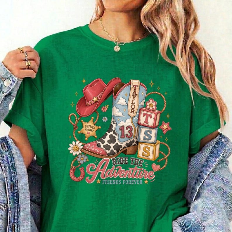 K&ouml;nnte beinhalten: Ein gr&uuml;nes T-Shirt mit einem Grafikdruck eines rosa Cowboyhuts, einem Cowboystiefel mit Kuhfleckenmuster und der Zahl 13 sowie gestapelten Bl&ouml;cken mit den Buchstaben T, S und 13. Das Design enth&auml;lt den Text Taylor, Adventure und Friends Forever.