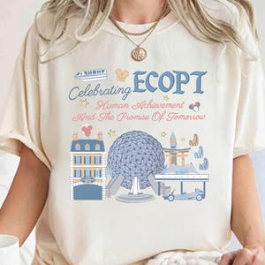 May include: Cream-colored t-shirt featuring a colorful graphic design. The design includes the word "ECOPT" in blue, along with the phrase "Celebrating Human Achievement And The Promise Of Tomorrow." The graphic also depicts various architectural landmarks.