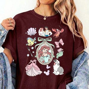 May include: A maroon t-shirt featuring a central oval frame containing an illustration of a red-haired character, surrounded by various icons including a seashell, a fork, a pink bow, a dog, a seahorse, a crab, and a pink dress.