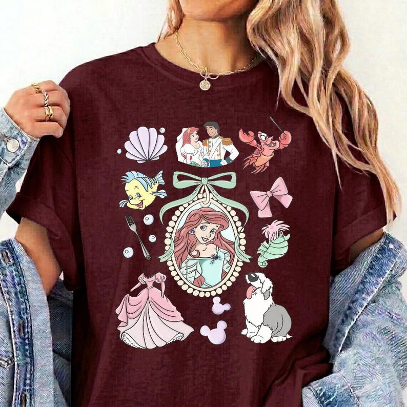 May include: A maroon t-shirt featuring a central oval frame containing an illustration of a red-haired character, surrounded by various icons including a seashell, a fork, a pink bow, a dog, a seahorse, a crab, and a pink dress.