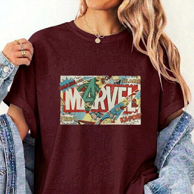 Puede incluir: Una camiseta de manga corta color granate con un estampado gr&aacute;fico rectangular en el pecho. El gr&aacute;fico muestra la palabra MARVEL en letras blancas grandes, superpuesta con paneles de c&oacute;mics antiguos que muestran personajes.
