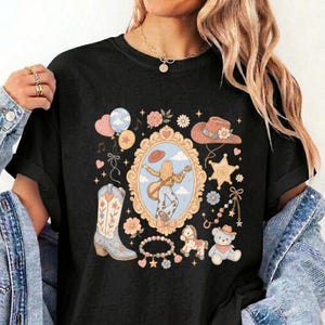 May include: A black t-shirt featuring a central graphic of a person playing a guitar inside an ornate gold frame. Surrounding the frame are illustrations of a cowboy boot, a teddy bear, a rocking horse, a cowboy hat, balloons, and flowers.
