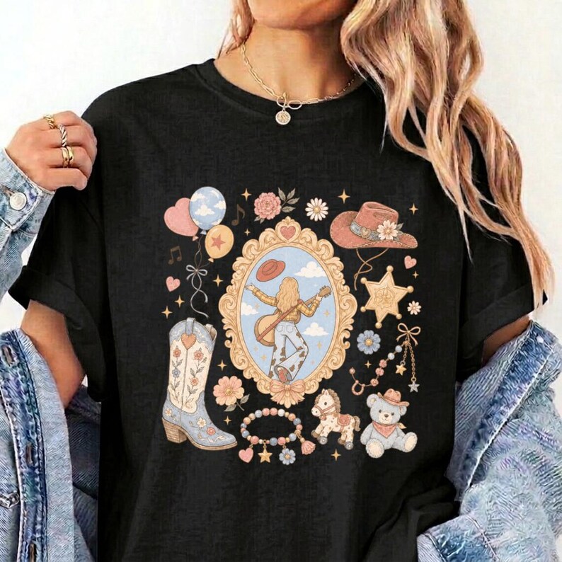 May include: A black t-shirt featuring a central graphic of a person playing a guitar inside an ornate gold frame. Surrounding the frame are illustrations of a cowboy boot, a teddy bear, a rocking horse, a cowboy hat, balloons, and flowers.