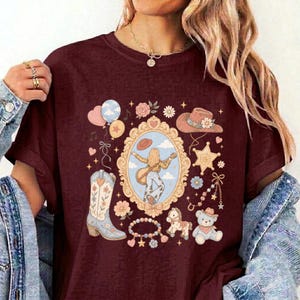 May include: A maroon t-shirt featuring a central ornate gold frame containing an illustration of a person playing a guitar. Surrounding the frame are graphics of a cowboy boot, a hat, a teddy bear, a rocking horse, balloons, and flowers.