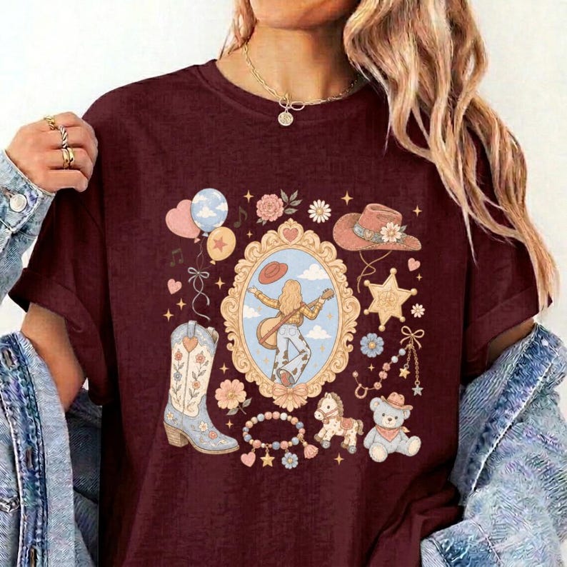 May include: A maroon t-shirt featuring a central ornate gold frame containing an illustration of a person playing a guitar. Surrounding the frame are graphics of a cowboy boot, a hat, a teddy bear, a rocking horse, balloons, and flowers.