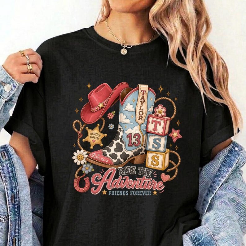 K&ouml;nnte beinhalten: Ein schwarzes T-Shirt mit einem Grafikdruck, der einen rosa Cowboyhut, einen Cowboystiefel mit Kuhfleckenmuster und der Zahl 13 sowie drei gestapelte Bl&ouml;cke mit den Buchstaben T, S und 13 zeigt. Das Design enth&auml;lt Sterne, Blumen, Seile und den Text Ride the Adventure Friends Forever.
