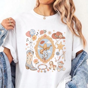 May include: A white t-shirt featuring a central graphic print of a person playing a guitar inside an ornate gold frame, surrounded by illustrations of a cowboy hat, a western boot, a teddy bear, balloons, flowers, and a star-shaped badge.