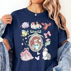 May include: A navy blue t-shirt featuring a central oval frame containing a red-haired character, surrounded by various icons including a seashell, a fork, a pink dress, a crab, a bow, a seahorse, a dog, and a couple in a wedding outfit.