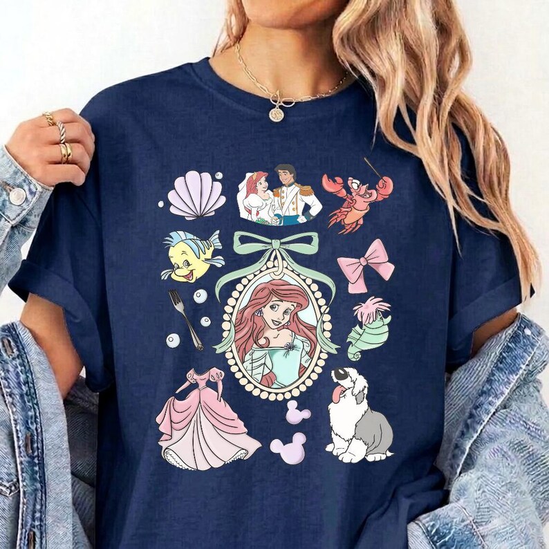May include: A navy blue t-shirt featuring a central oval frame containing a red-haired character, surrounded by various icons including a seashell, a fork, a pink dress, a crab, a bow, a seahorse, a dog, and a couple in a wedding outfit.