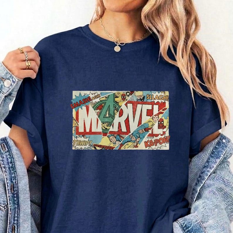 Puede incluir: Una camiseta azul marino con un estampado gr&aacute;fico rectangular de una portada de c&oacute;mic vintage. El dise&ntilde;o muestra la palabra MARVEL en letras rojas grandes, superpuesta con paneles de c&oacute;mic, figuras de acci&oacute;n y onomatopeyas como SMASH.