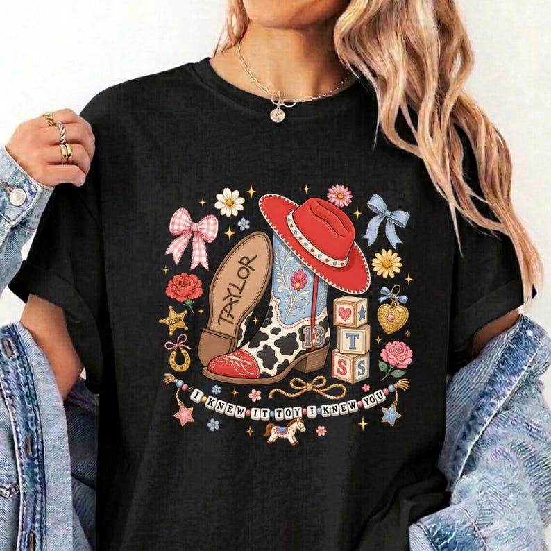 May include: A black t-shirt featuring a graphic print of a red cowboy hat, a cow-print boot with the name Taylor, and alphabet blocks spelling TS. The design includes pink bows, flowers, stars, and the text I KNEW IT TOY I KNEW YOU.
