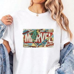 Puede incluir: Una camiseta blanca de manga corta con un estampado gr&aacute;fico rectangular central. El dise&ntilde;o muestra la palabra MARVEL en letras rojas grandes, superpuesta sobre un collage de paneles de c&oacute;mics antiguos con palabras de acci&oacute;n.