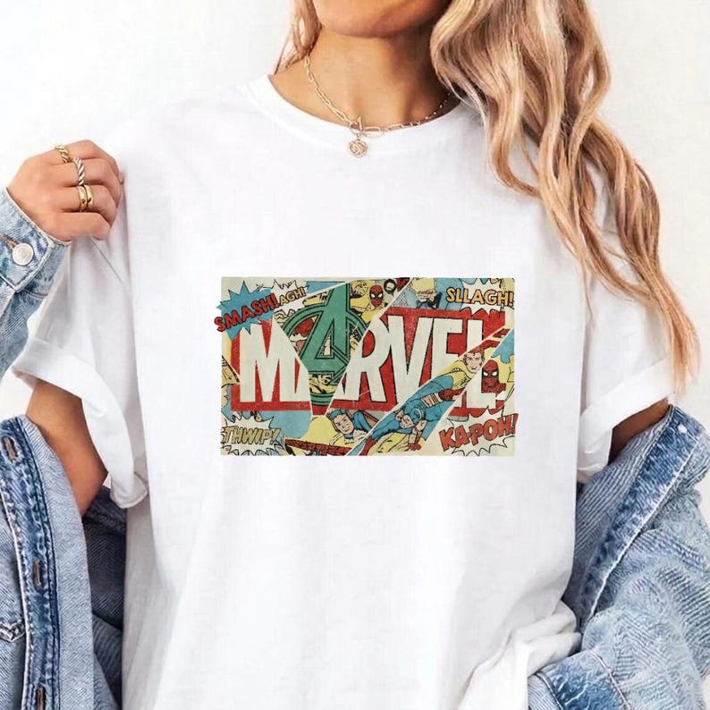 Puede incluir: Una camiseta blanca de manga corta con un estampado gr&aacute;fico rectangular central. El dise&ntilde;o muestra la palabra MARVEL en letras rojas grandes, superpuesta sobre un collage de paneles de c&oacute;mics antiguos con palabras de acci&oacute;n.