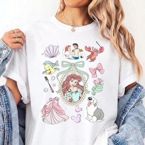 May include: A white t-shirt featuring a colourful graphic print of various characters and items from The Little Mermaid, including Ariel in a central oval frame, a seashell, a fork, a pink dress, a crab, a seahorse, a dog, and a bow.