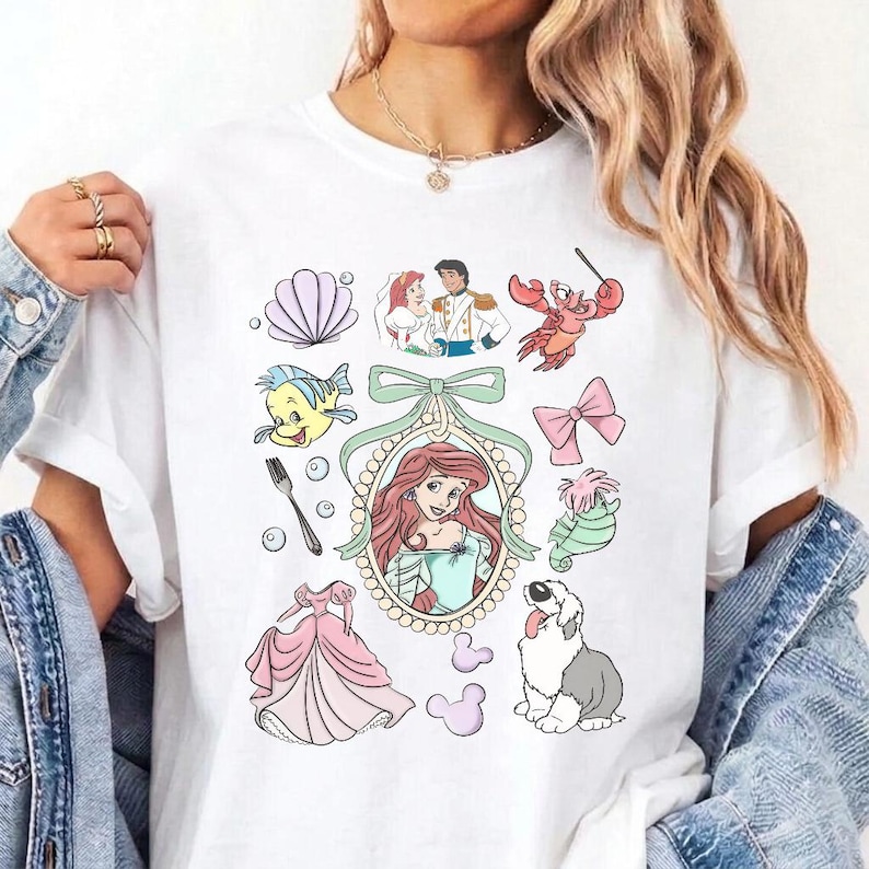 May include: A white t-shirt featuring a colourful graphic print of various characters and items from The Little Mermaid, including Ariel in a central oval frame, a seashell, a fork, a pink dress, a crab, a seahorse, a dog, and a bow.