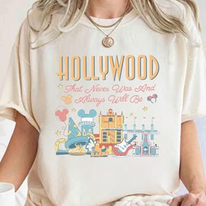 May include: Cream-colored t-shirt with a colorful graphic design featuring the word "HOLLYWOOD" in orange and blue, along with the phrase "That Never Was And Always Will Be." The design includes cartoon illustrations of iconic landmarks.
