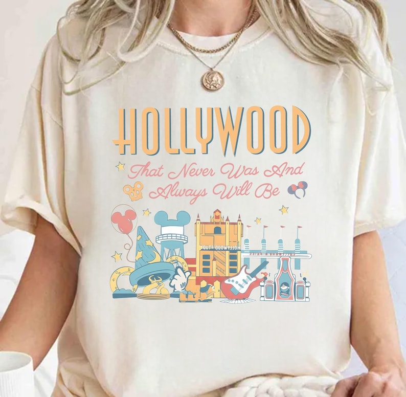 May include: Cream-colored t-shirt with a colorful graphic design featuring the word "HOLLYWOOD" in orange and blue, along with the phrase "That Never Was And Always Will Be." The design includes cartoon illustrations of iconic landmarks.