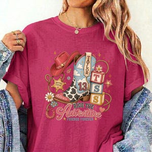 K&ouml;nnte beinhalten: Ein magentafarbenes T-Shirt mit einem Grafikdruck eines Cowboystiefels mit Kuhfleckenmuster, einem rosa Cowboyhut und drei gestapelten Bl&ouml;cken mit der Aufschrift 13, T und S. Das Design enth&auml;lt Sterne, Blumen und den Text Ride the Adventure Friends Forever.