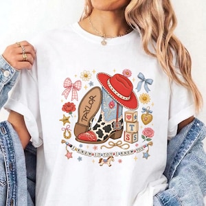 May include: A white t-shirt featuring a graphic print of a red cowboy hat, a cow-print boot with the number 13, a wooden block stack, and the text TAYLOR and I KNEW IT TOY I KNEW YOU, surrounded by illustrations of bows, flowers, and a rocking horse.