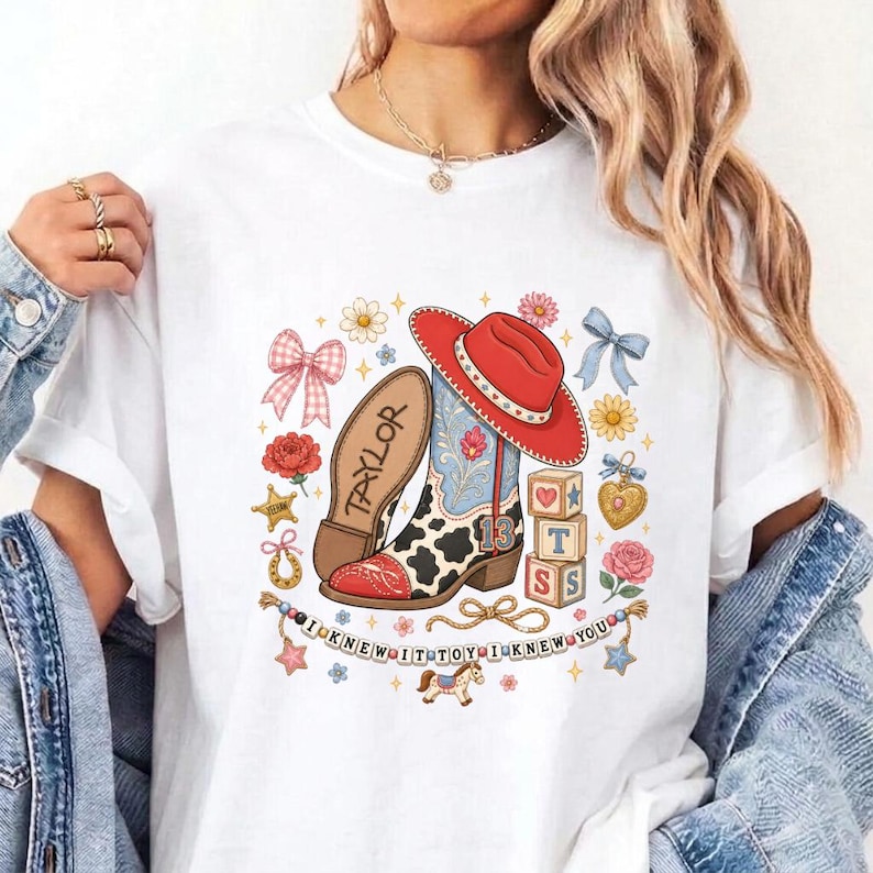 May include: A white t-shirt featuring a graphic print of a red cowboy hat, a cow-print boot with the number 13, a wooden block stack, and the text TAYLOR and I KNEW IT TOY I KNEW YOU, surrounded by illustrations of bows, flowers, and a rocking horse.