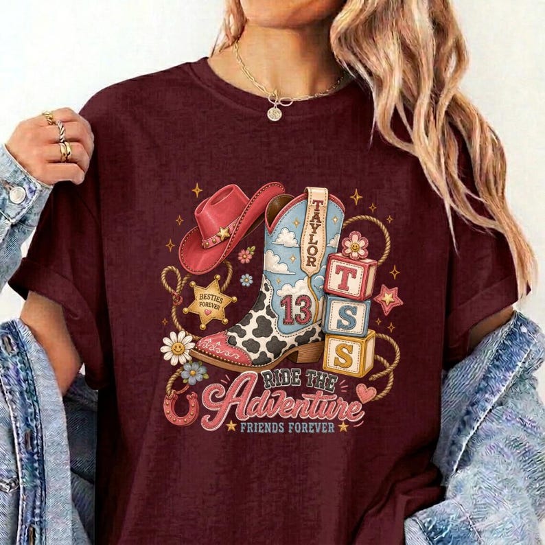 K&ouml;nnte beinhalten: Ein kastanienbraunes T-Shirt mit einem Grafikdruck eines rosa Cowboyhuts, eines Cowboystiefels mit Kuhfleckenmuster und der Zahl 13 sowie drei gestapelten Bl&ouml;cken mit den Buchstaben T, S und 13. Der Text lautet Taylor, Ride the Adventure, Besties Forever.