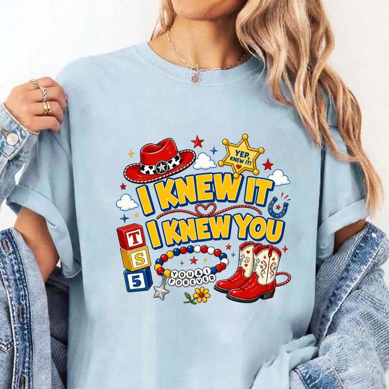 Puede incluir: Una camiseta azul claro con un estampado gr&aacute;fico que incluye un sombrero de vaquero rojo, una estrella de sheriff amarilla con el texto YEP KNEW IT, botas de vaquero rojas y blancas, bloques de letras y una pulsera con cuentas.